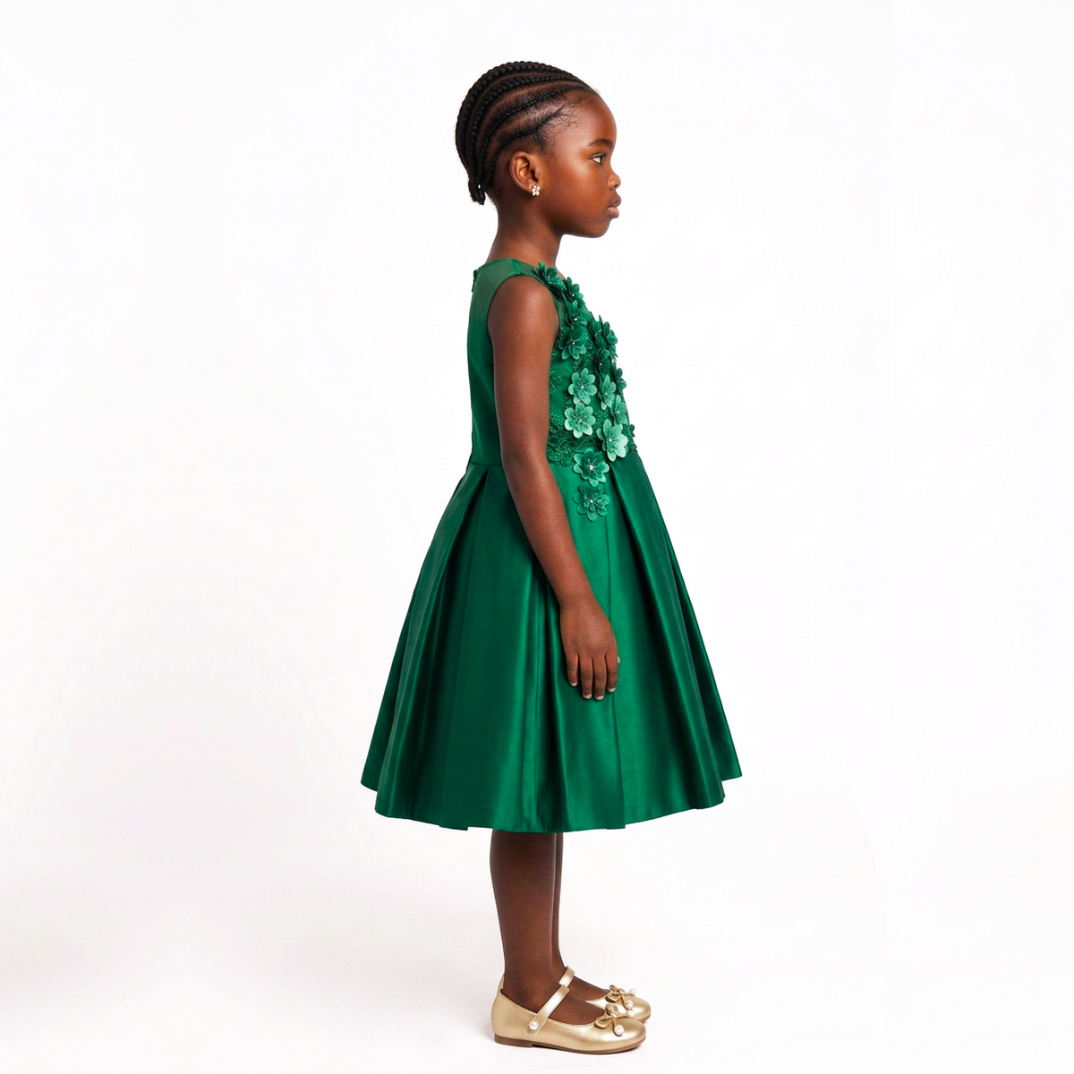 A young girl with braided hair is shown in profile wearing a sleeveless emerald green dress with 3D floral applique on the bodice and gold flat shoes, photographed against a solid white studio background.