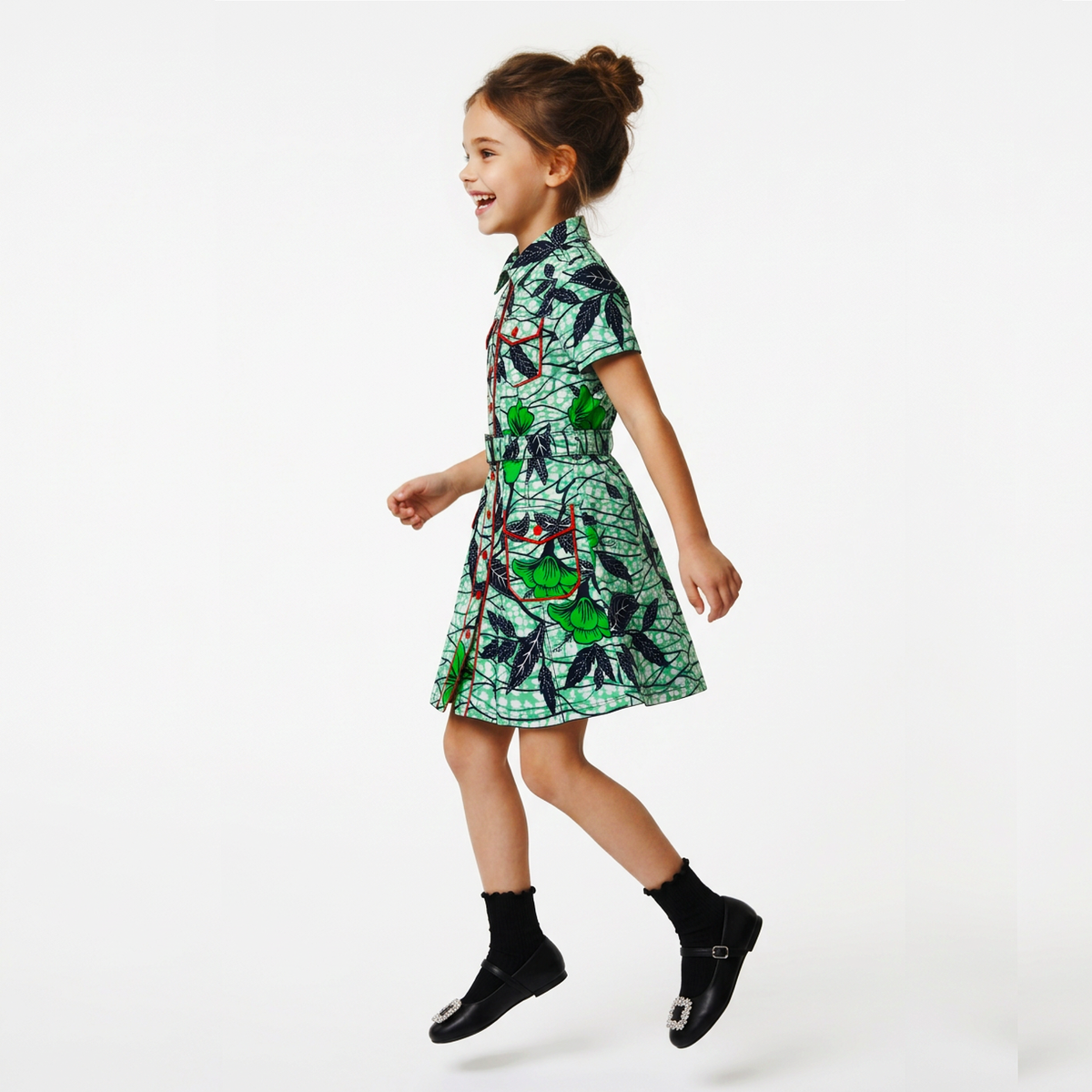 A young girl with her hair in a bun is shown from the side, wearing a short-sleeved green dress with a black and red abstract pattern, paired with black socks and black shoes, against a plain white background.
