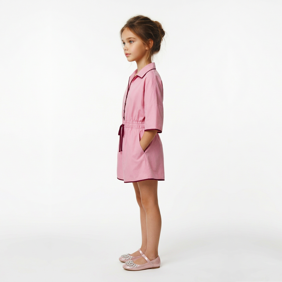 A young girl stands in profile wearing a pink short-sleeve collared dress with a tie waist and side pockets, paired with light pink embellished flats, against a plain white studio background.