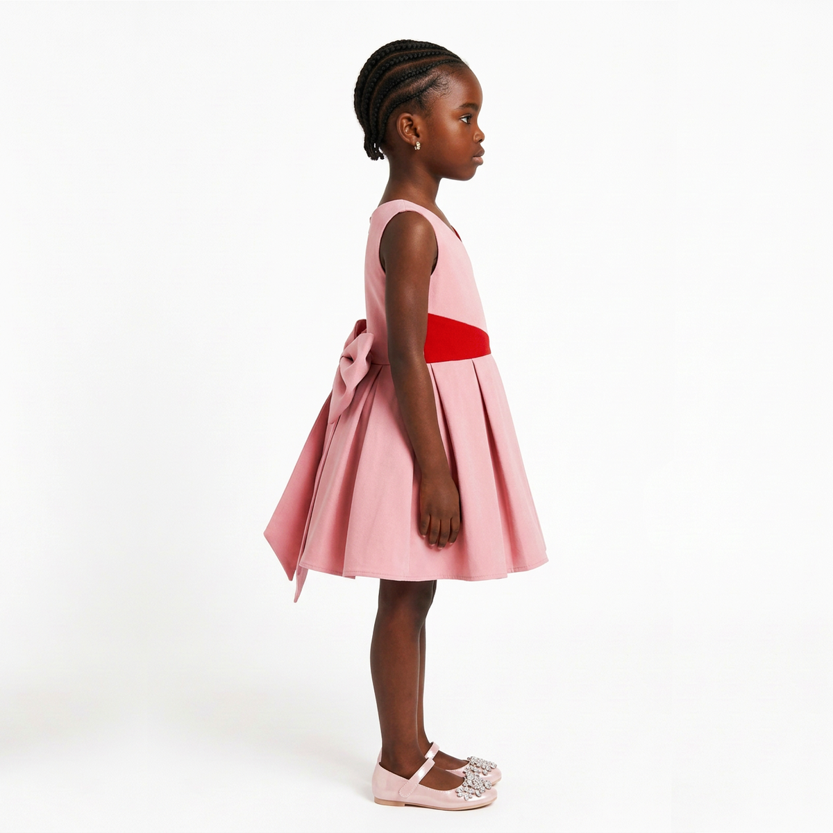 A young girl with braided hair is shown in profile wearing a sleeveless pink dress with a red waist sash and a large bow at the back of the skirt. She is standing against a clean white studio background.