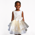 A young girl with braided hair is wearing a sleeveless white dress with a large white bow at the waist and a layered tulle skirt in white and pale yellow. She is standing against a plain white studio background.