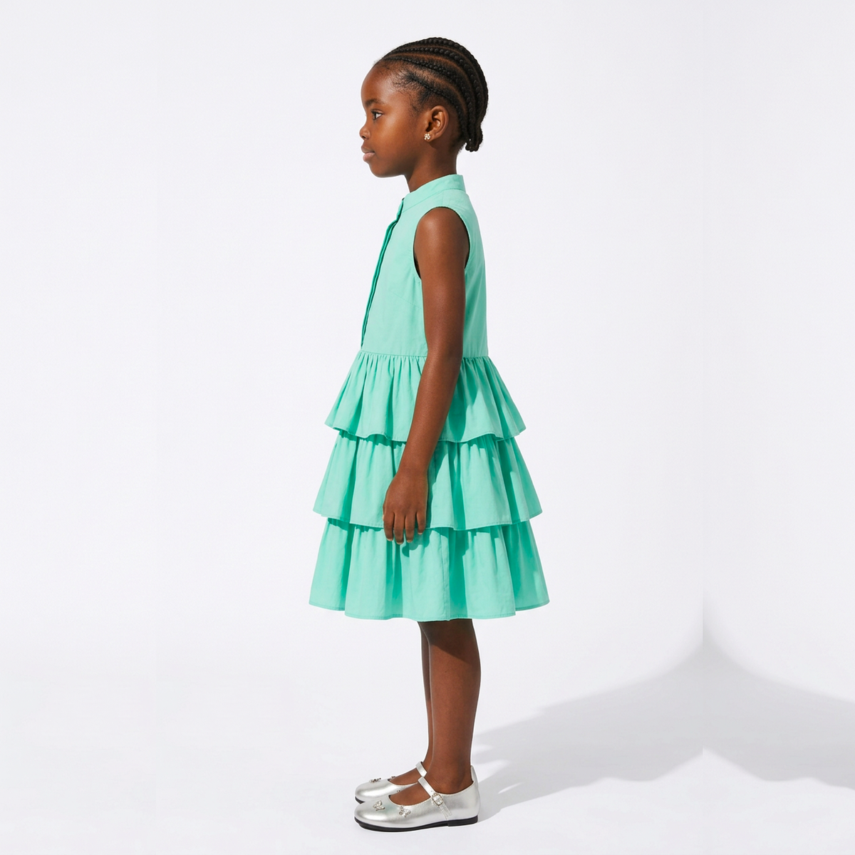 A young girl in profile wearing a sleeveless tiered dress in mint green, paired with silver Mary Jane shoes, standing against a clean white studio background.