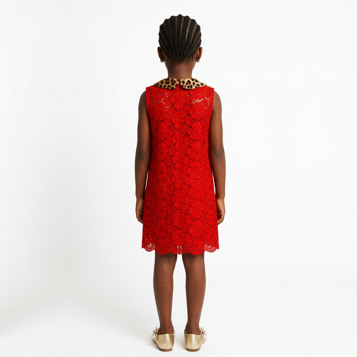 A young girl viewed from behind wearing a sleeveless bright red lace dress with a leopard-print collar and gold flat shoes, photographed against a plain white studio background.