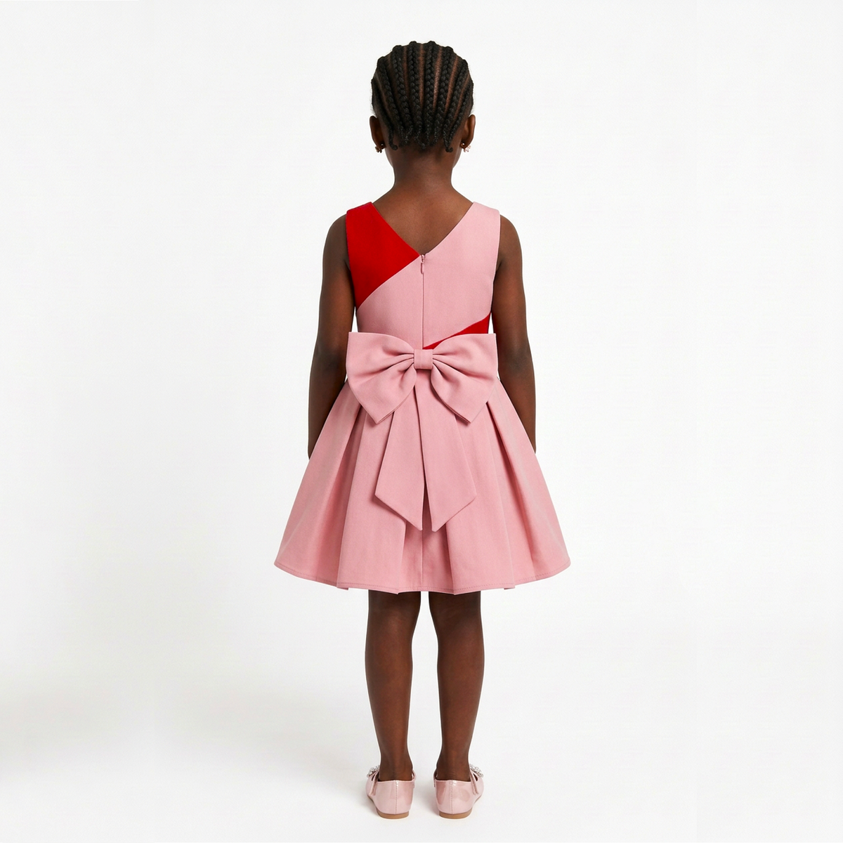 A young girl viewed from behind wearing a sleeveless pink dress with a red yoke detail and a large bow at the back, standing against a plain white studio background.