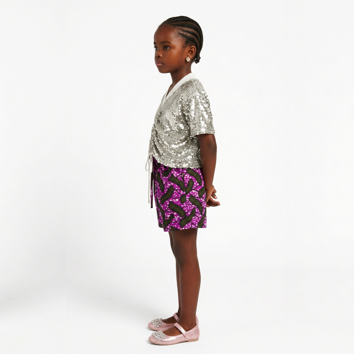 A young girl standing in profile wearing a silver sequined wrap-style top and purple shorts with a black floral pattern, paired with pink flats, against a plain white studio background.