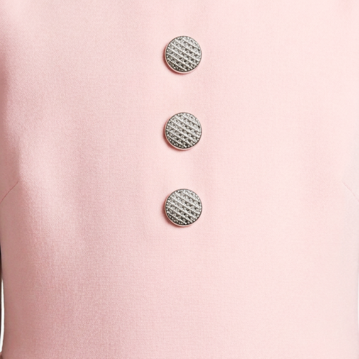 A close-up of a light pink garment featuring three vertically aligned round silver buttons with a textured, woven-like pattern.