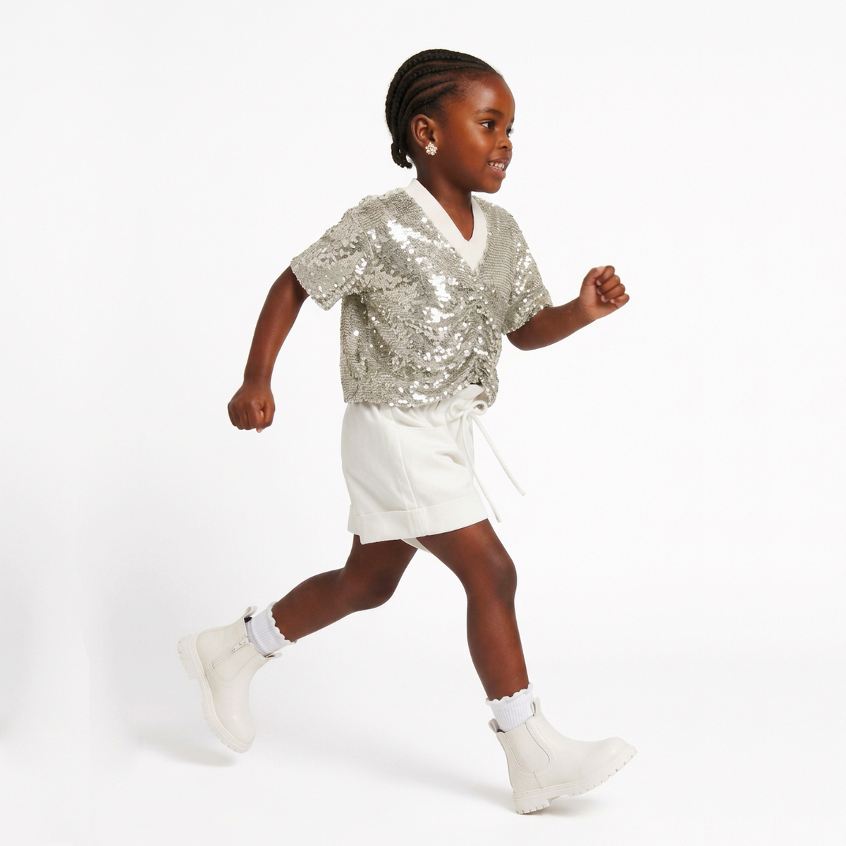 A young girl running in a studio wearing a silver sequined short-sleeve wrap top, white shorts, and white boots against a plain white background.