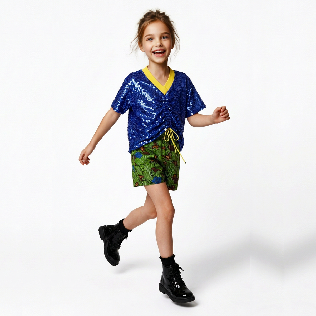 A young girl smiling and running while wearing a blue sequined V-neck t-shirt with a yellow trim, green patterned shorts, and black boots against a white studio background.
