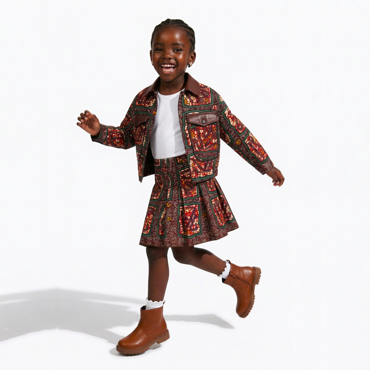 A young girl smiling and walking in a studio while wearing a matching brown and red patterned jacket and pleated skirt set, a white t-shirt, white socks, and brown boots against a white background.