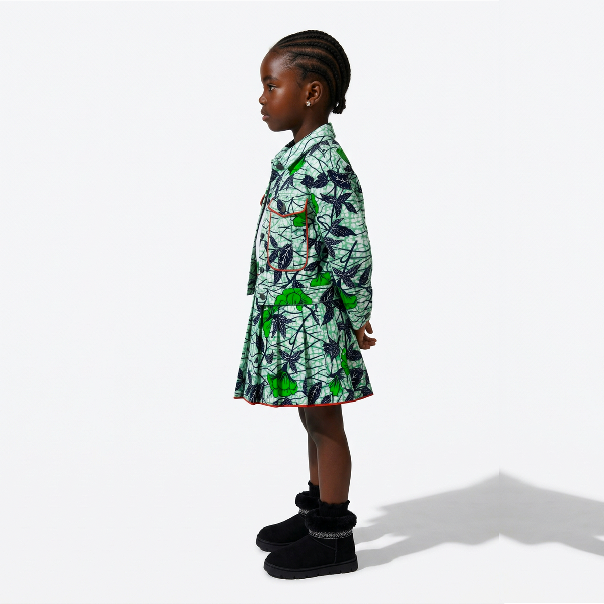 A young child stands in profile wearing a short green and black patterned dress with bright green accents, paired with black boots, against a clean white studio background.