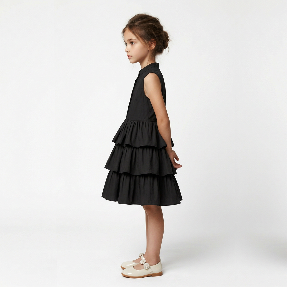 A young girl with her hair in a bun stands in profile wearing a sleeveless black tiered dress and cream Mary Jane shoes against a plain white studio background.