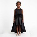 A young girl stands against a plain white studio background wearing a sleeveless black dress with a feathered waist detail and a high-low skirt, paired with white shoes.