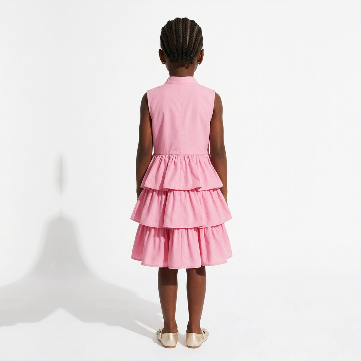 A young girl viewed from behind wearing a sleeveless pink dress with a tiered ruffle skirt, standing against a plain white studio background.
