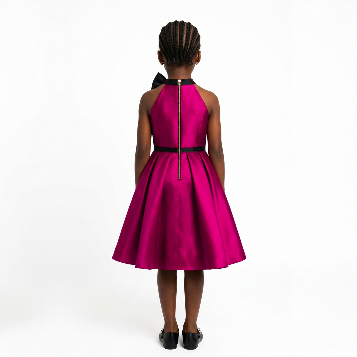 A young girl viewed from behind wearing a bright magenta pink halter dress with black trim, a black bow at the back of the neck, and a black waistband, paired with black shoes, against a plain white background.