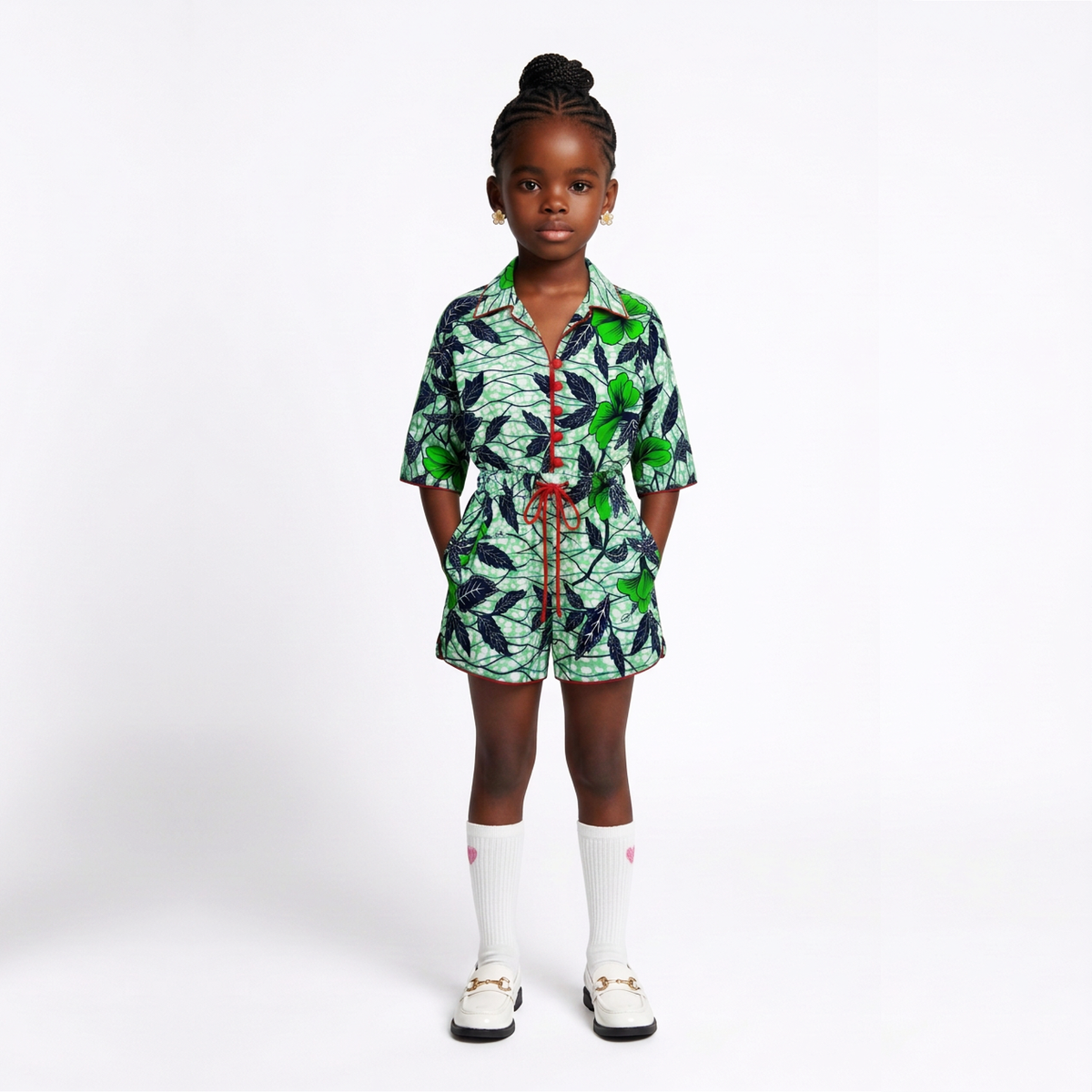 A young child stands against a light gray studio background wearing a short-sleeve button-front shirt and matching shorts set in a green, black, and white leaf print, with white knee-high socks and white shoes.