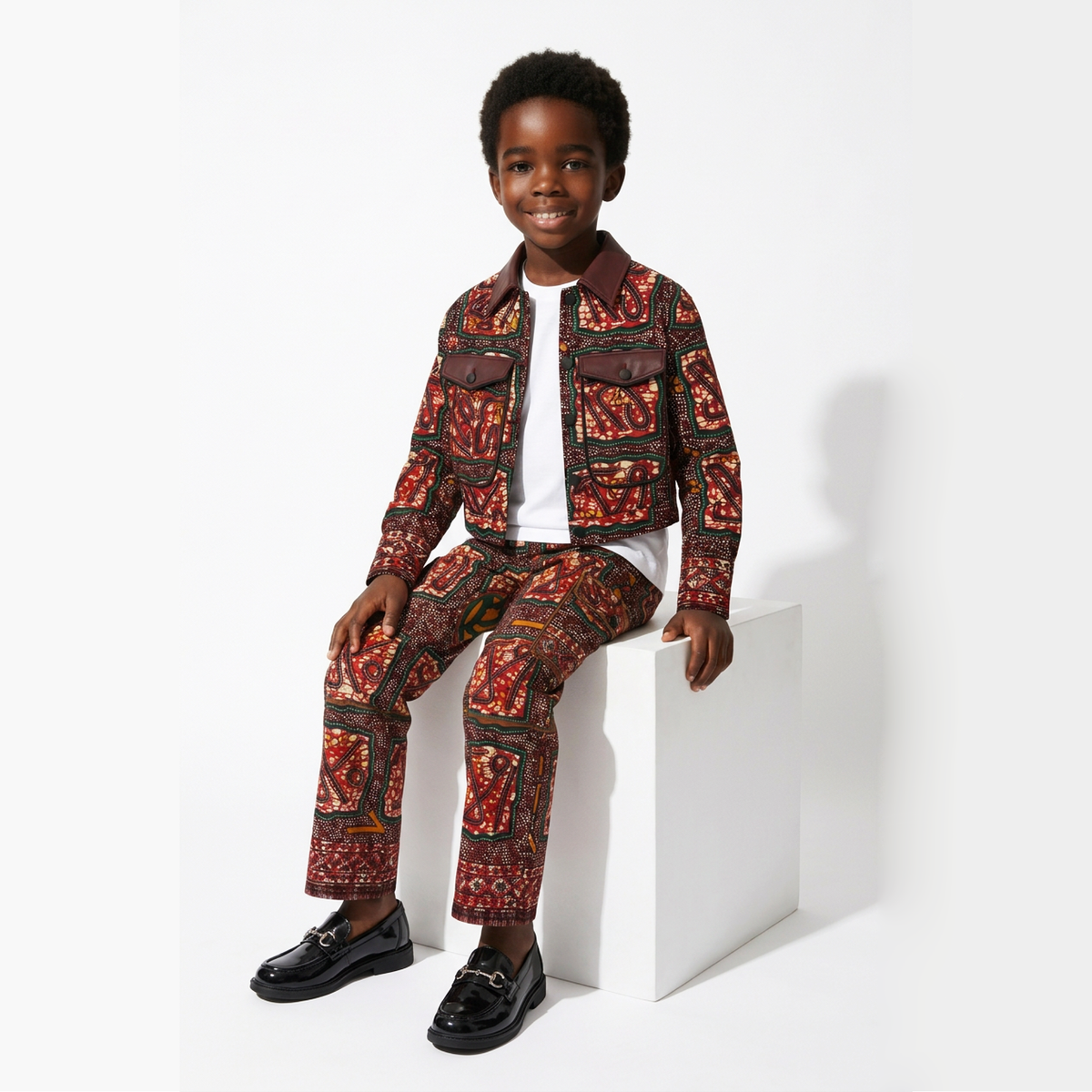 A young boy models a matching two-piece suit featuring a red, brown, and black traditional African wax print pattern, styled with a white t-shirt, black loafers, and sits on a white block against a white studio background.