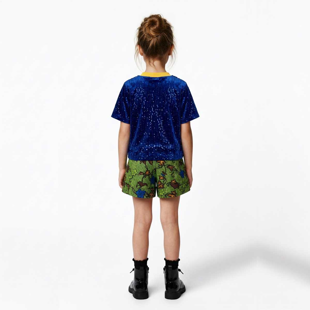 A child model shown from the back wearing a short-sleeve sequined t-shirt in blue and green, patterned shorts, and black boots, set against a plain white studio background.