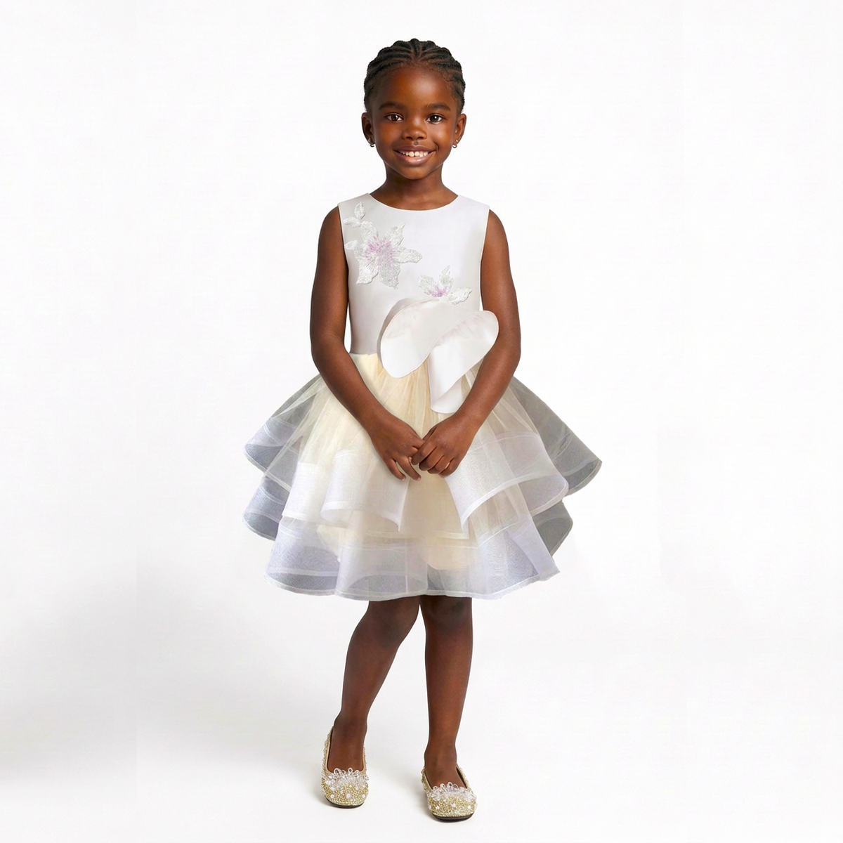 A young girl stands against a white studio background wearing a sleeveless white and cream tulle dress with floral applique on the bodice and a large bow at the waist. She is also wearing sparkly gold shoes.