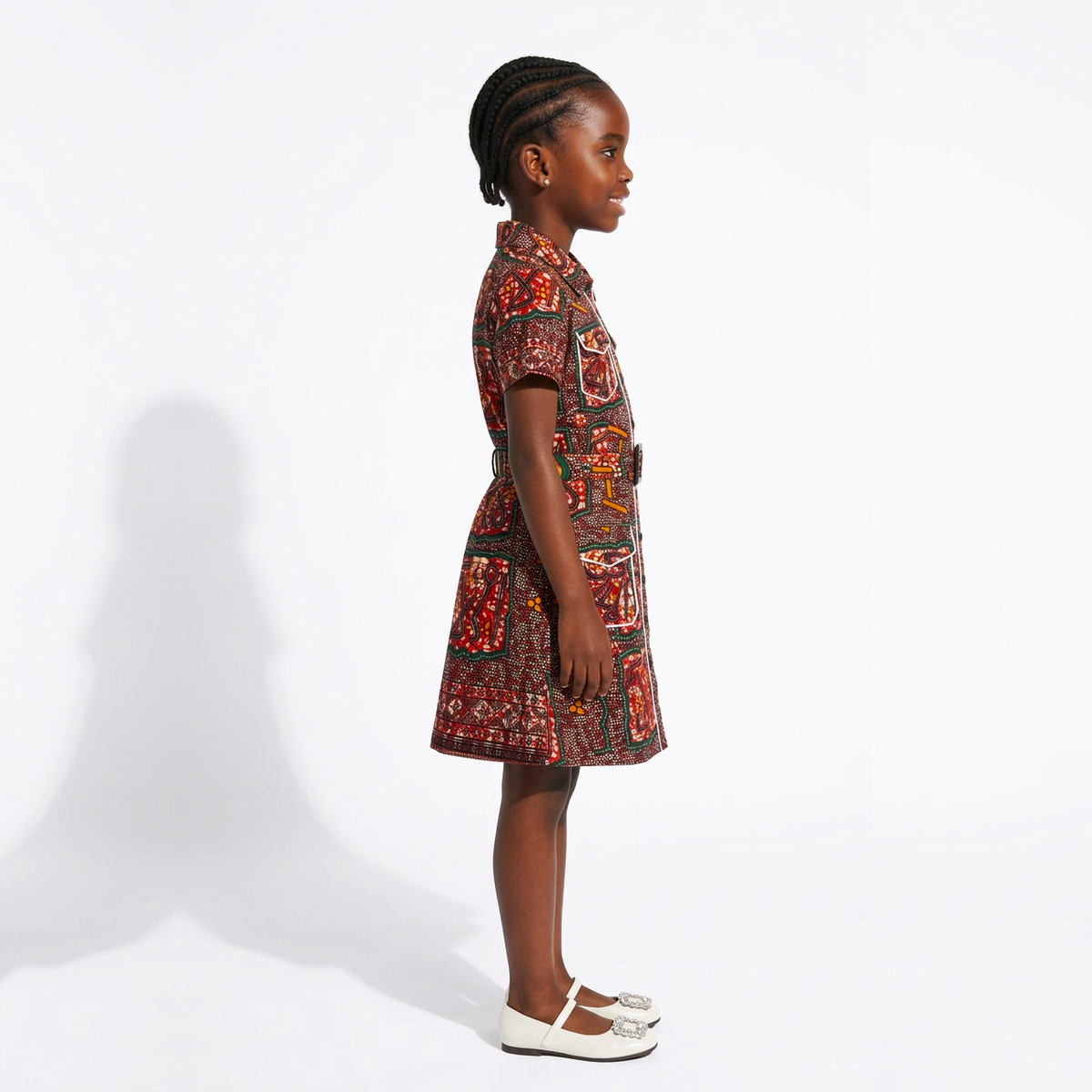 A young girl in profile wearing a short-sleeve collared dress with a red, brown, and black patterned print, paired with white Mary Jane shoes, standing against a white studio background.
