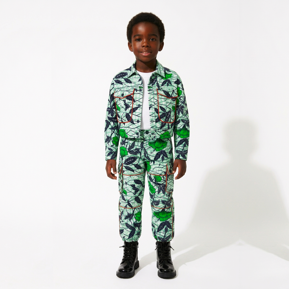A young boy stands against a white studio background wearing a matching long-sleeve button-front jacket and pants set with a bold tropical leaf print in green, black, and white, over a white undershirt and black boots.