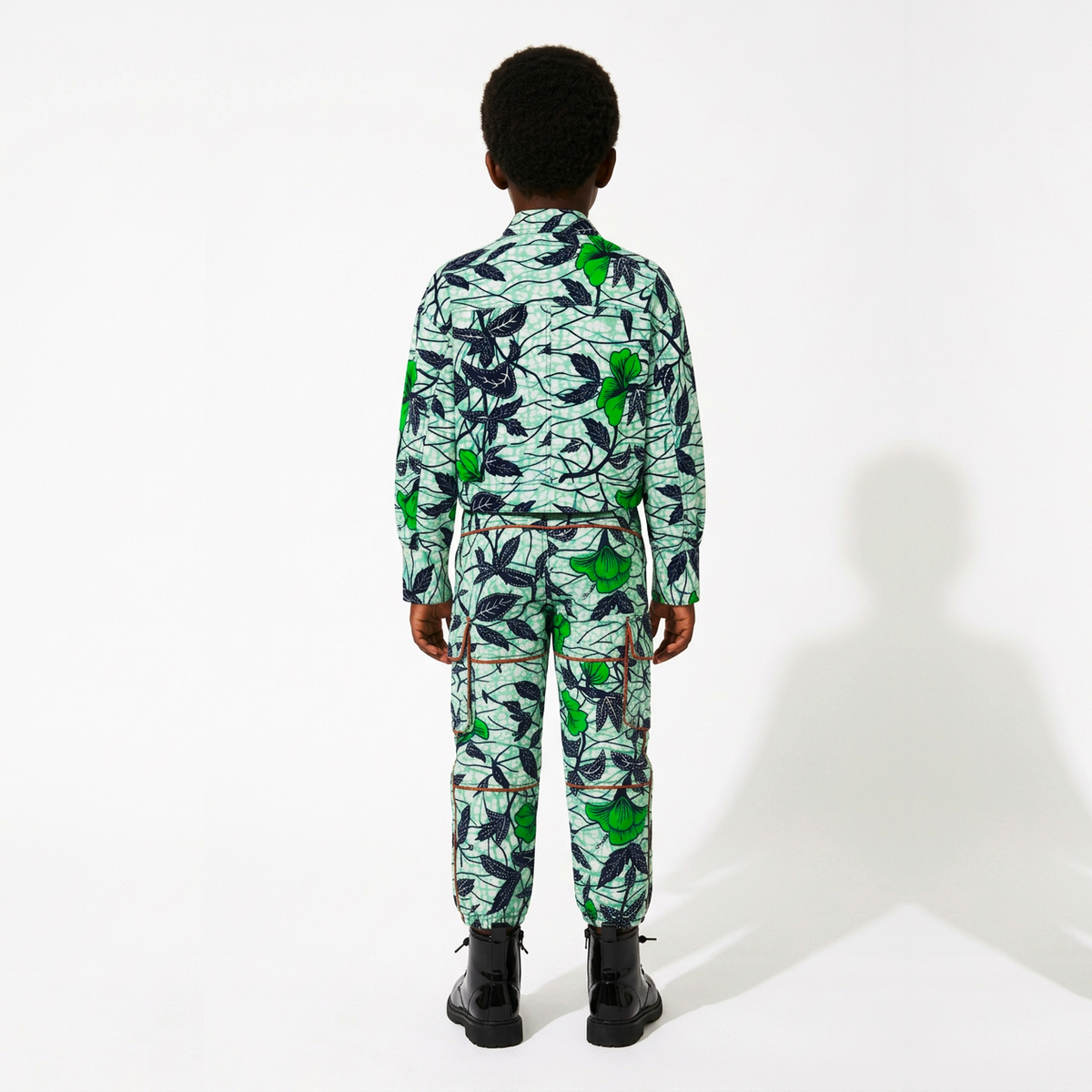 A child model viewed from the back wearing a matching long-sleeve collared shirt and pants set with a green and navy botanical print on a light mint background, paired with black boots, against a white studio background.