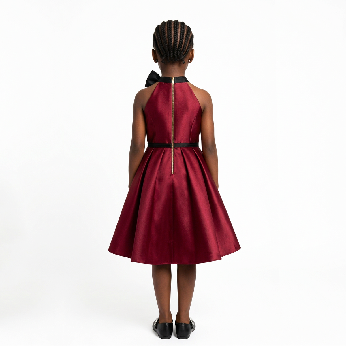A young girl viewed from behind wearing a sleeveless halter midi dress in deep burgundy with black trim, a black bow at the back of the neck, and a thin black waistband with a central gold zipper. She is standing against a plain white studio background.
