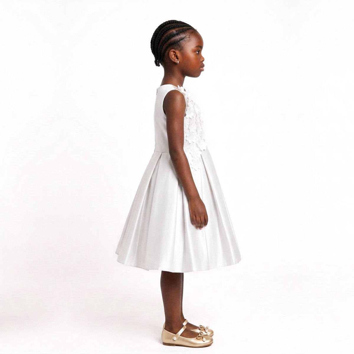 A young girl with braided hair stands in profile wearing a sleeveless white dress with lace detailing on the bodice and a flared skirt, paired with metallic gold shoes, against a plain white studio background.