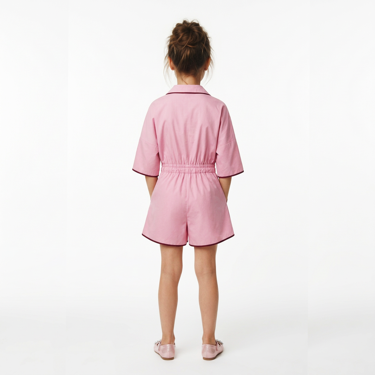 A young girl viewed from behind wearing a pink short-sleeve romper with dark pink piping, standing against a plain white studio background.