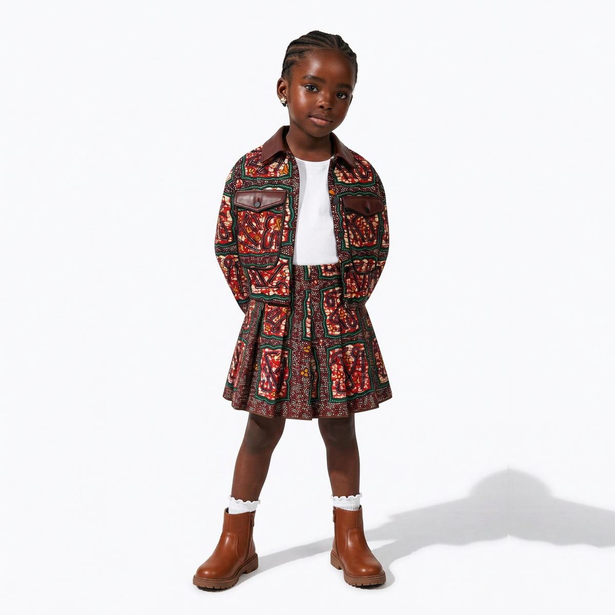 A young girl modeling a matching patterned jacket and pleated skirt set in a red, black, and gold colorway, styled with a white top and brown boots against a white studio background.