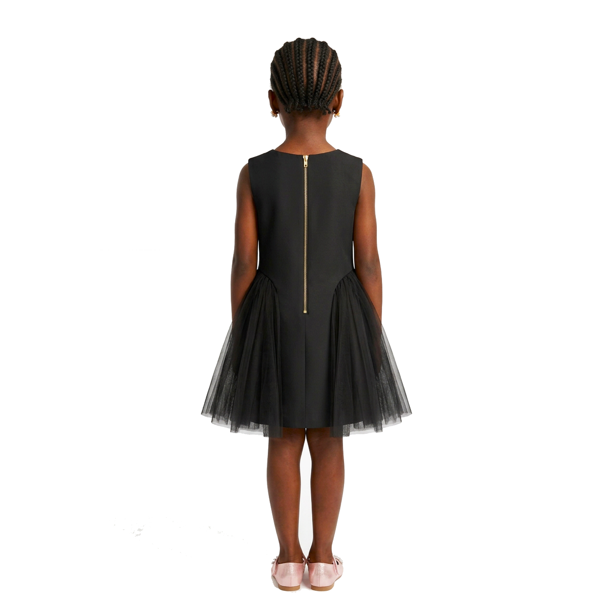 A young girl viewed from the back wearing a sleeveless black dress with a tulle skirt and a gold zipper detail at the back, set against a plain white background.