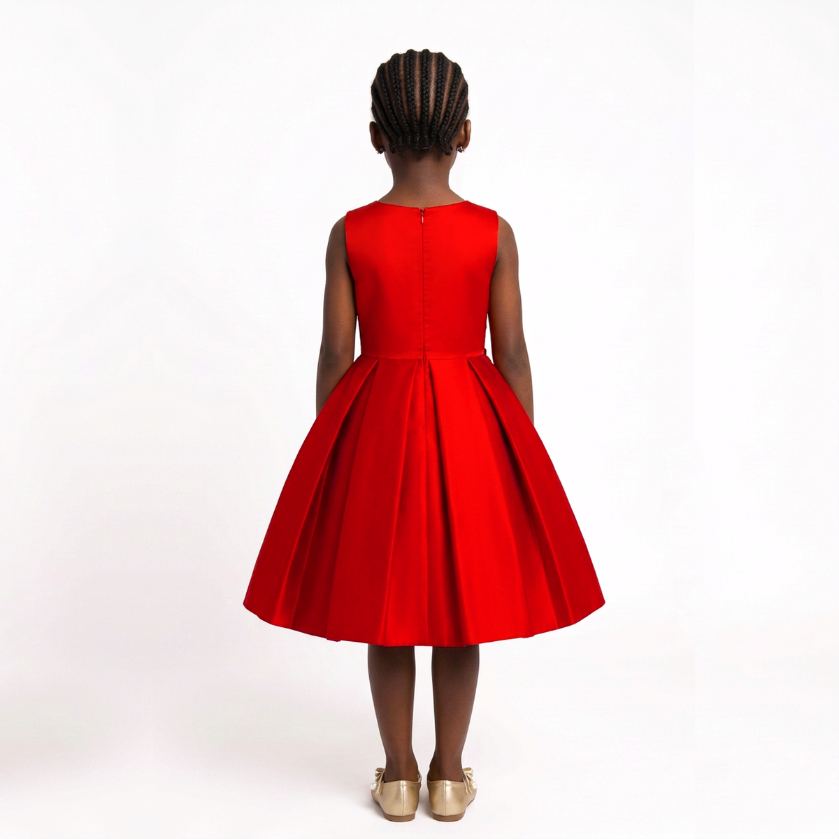 A young girl viewed from behind wearing a bright red sleeveless dress with a pleated skirt, paired with metallic gold shoes, set against a plain white studio background.