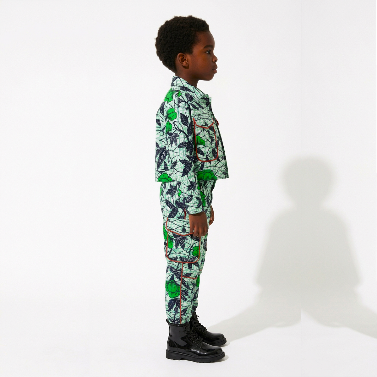 A young boy stands in profile wearing a matching long-sleeve button-up shirt and trousers set with a vibrant green tropical leaf print on a white base, paired with black boots, against a plain white studio background.
