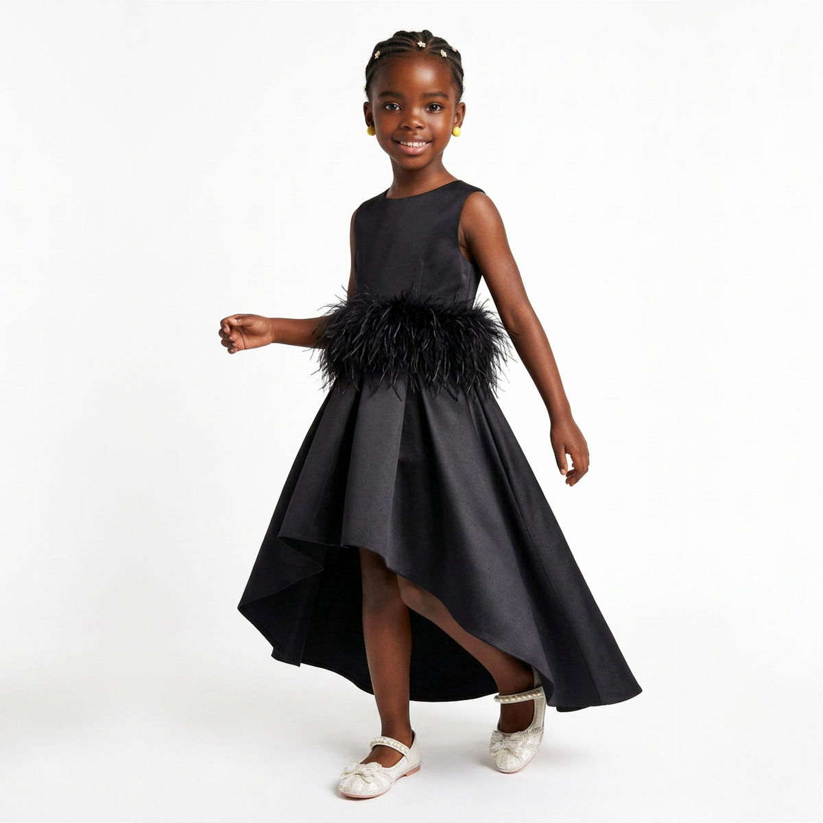 A young girl modeling a sleeveless black high-low dress with a feathered waistband, paired with white Mary Jane shoes, against a plain white studio background.