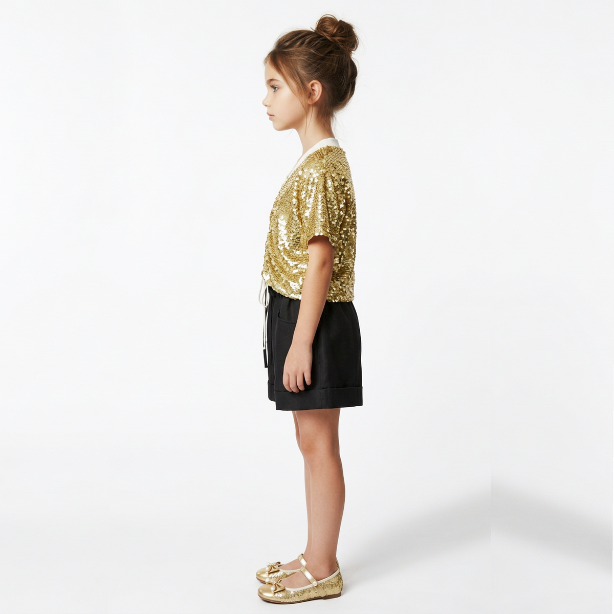 A young girl in profile wearing a gold sequined short-sleeve top, black shorts, and gold flat shoes, standing against a plain white studio background.