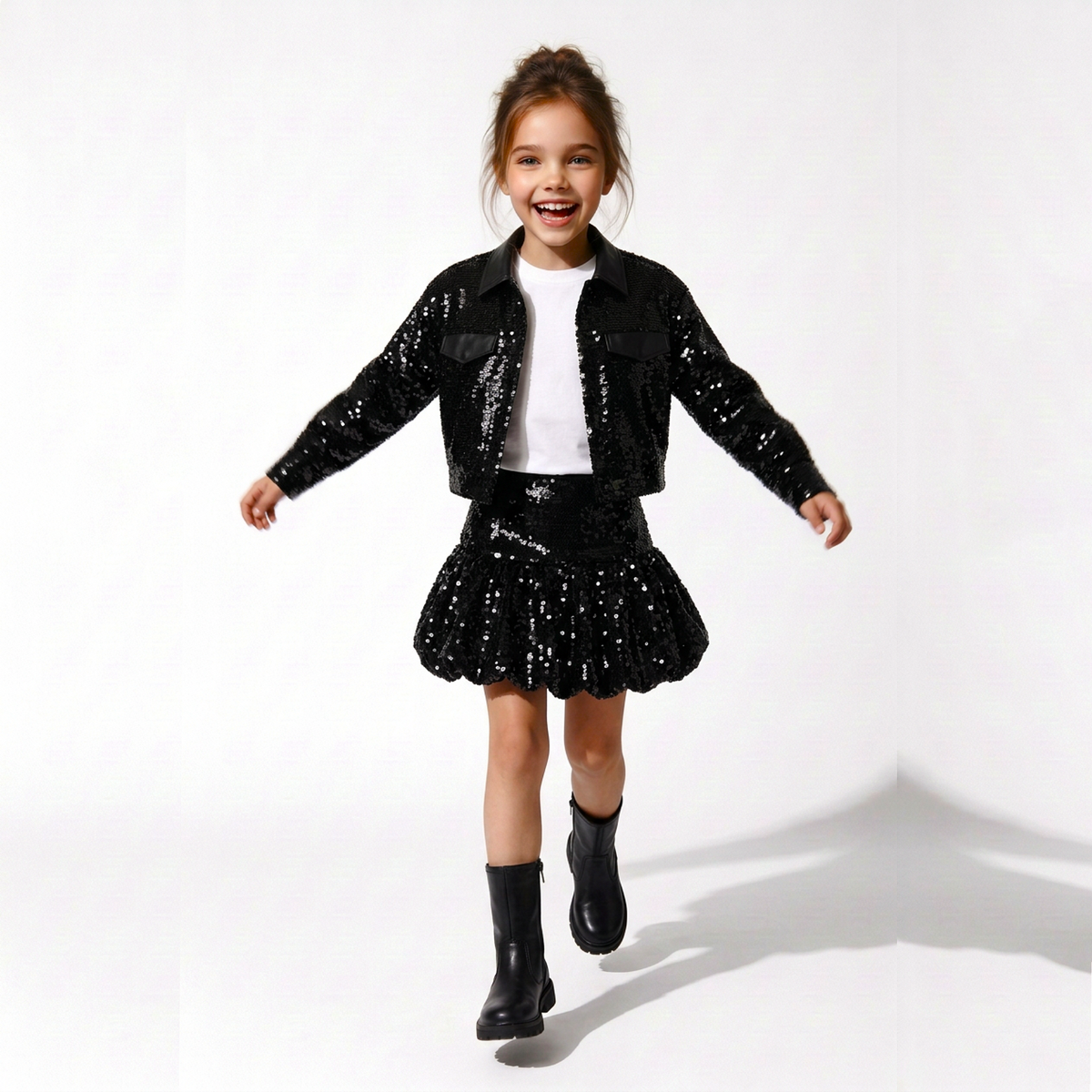 A young girl smiling and running while wearing a black sequined jacket, matching sequined mini skirt, white top, and black boots against a white studio background.