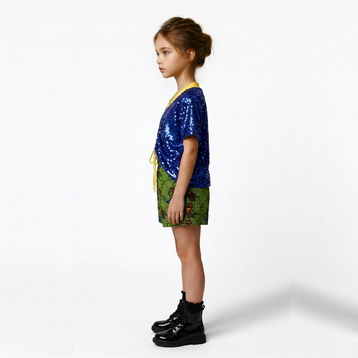 A young girl stands in profile wearing a short blue sequined top with yellow trim, green patterned shorts, and black boots against a plain white studio background.
