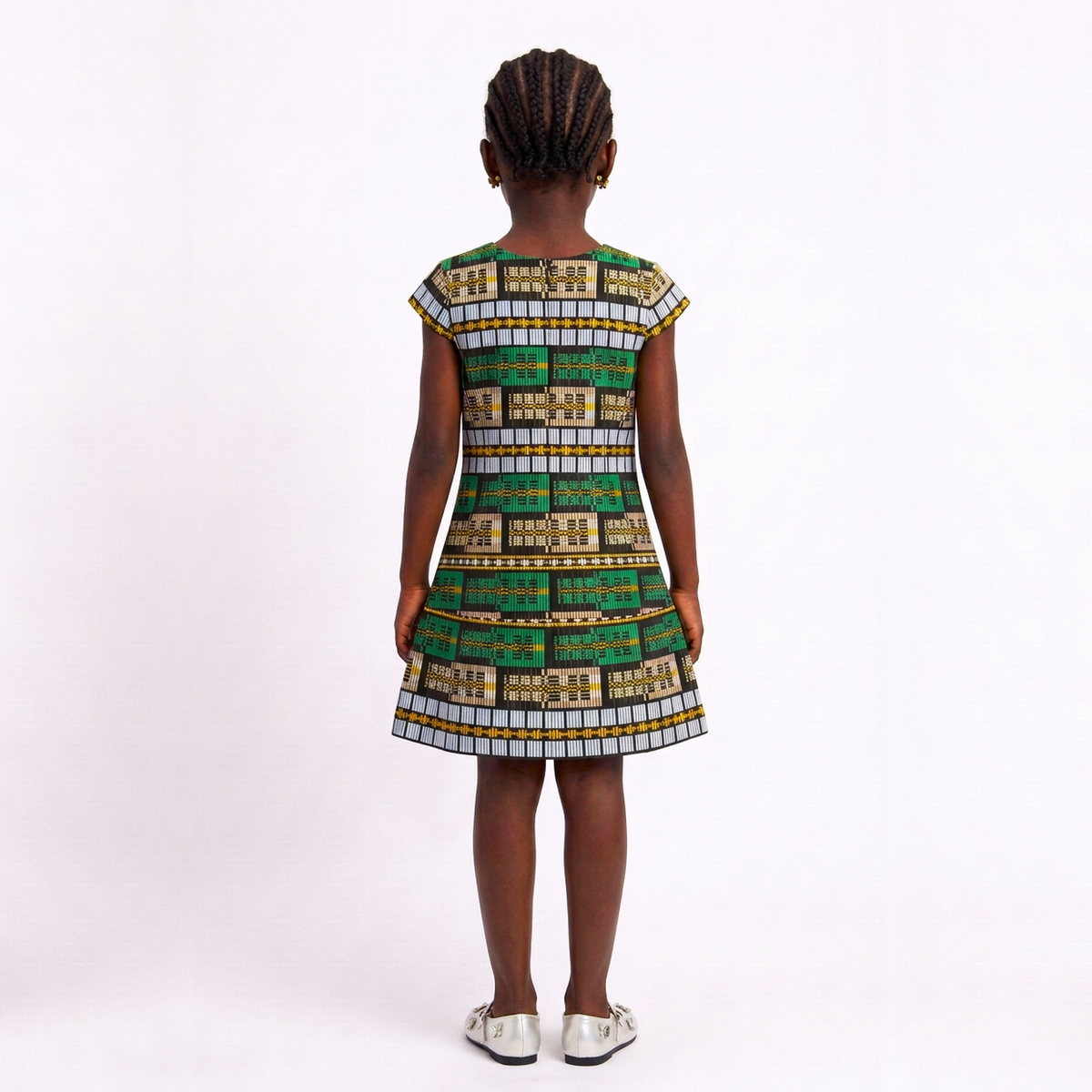 A young girl viewed from behind wearing a sleeveless A-line dress with a colorful geometric pattern in green, gold, and white, paired with white shoes, set against a plain white studio background.
