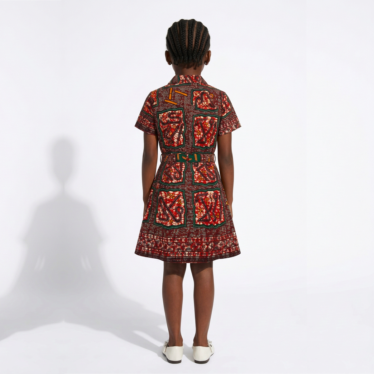 A child model shown from the back wearing a short-sleeve collared dress with an intricate red, orange, and black geometric pattern, paired with white shoes, against a white studio background.