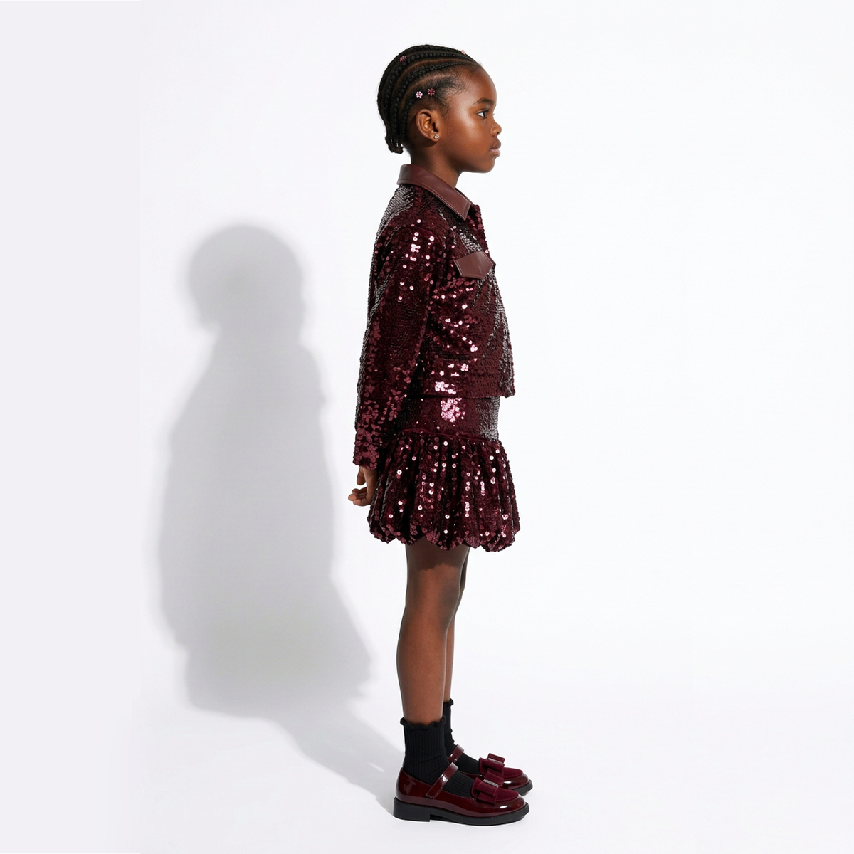 A young girl in profile wearing a sparkling burgundy sequined jacket and matching pleated skirt, paired with black socks and burgundy shoes, standing against a white studio background with a soft shadow.