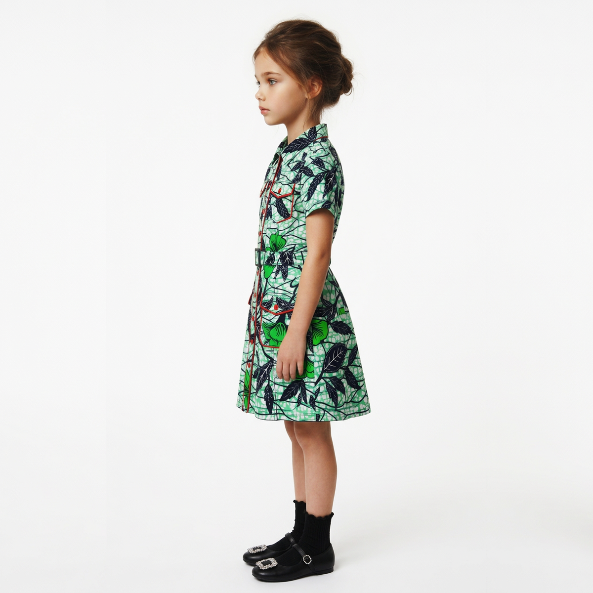 A young girl stands in profile wearing a short-sleeve collared dress with a green and black tropical leaf print, paired with black socks and black shoes, against a plain white studio background.