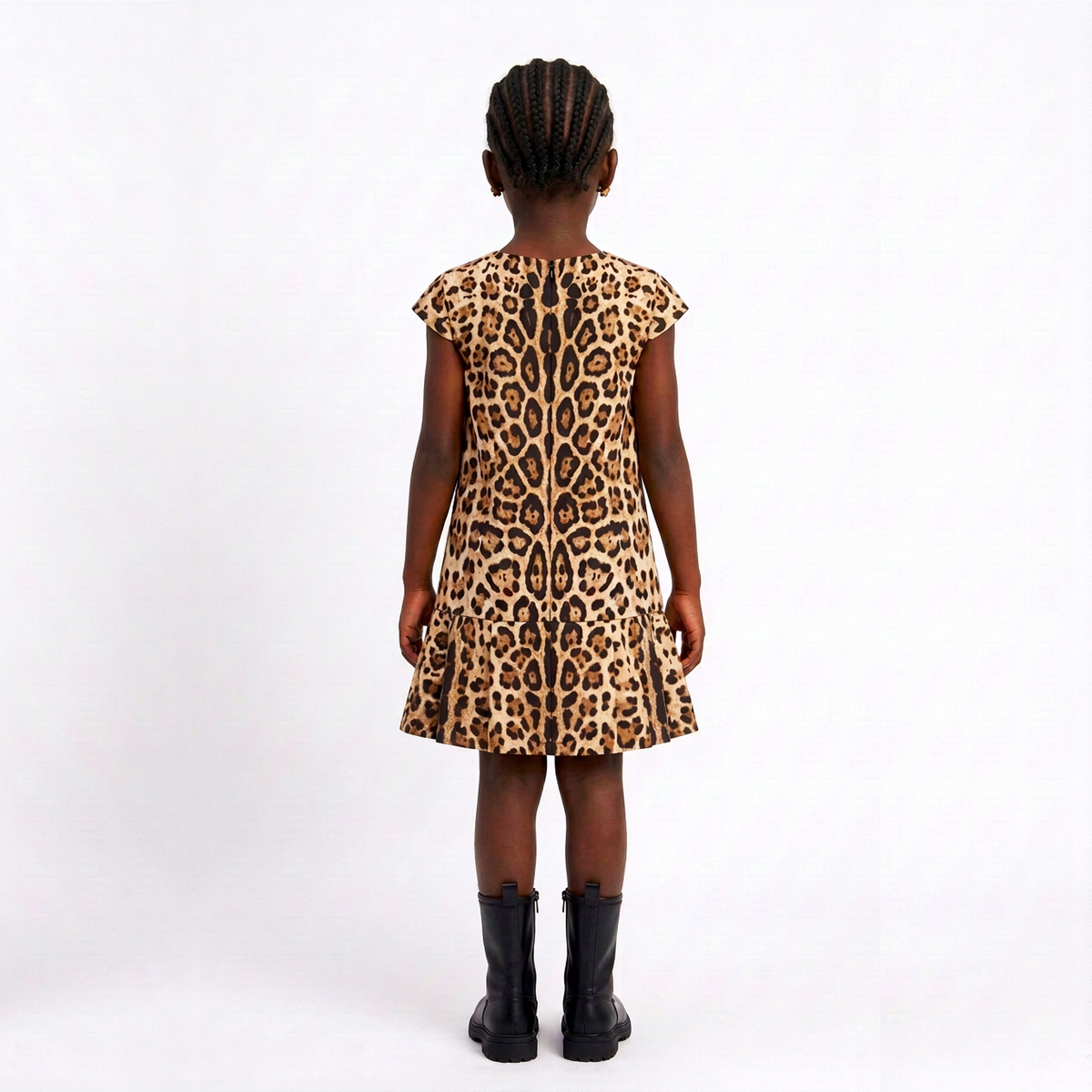 A young girl viewed from behind wearing a leopard print dress and black boots, standing against a plain white studio background.