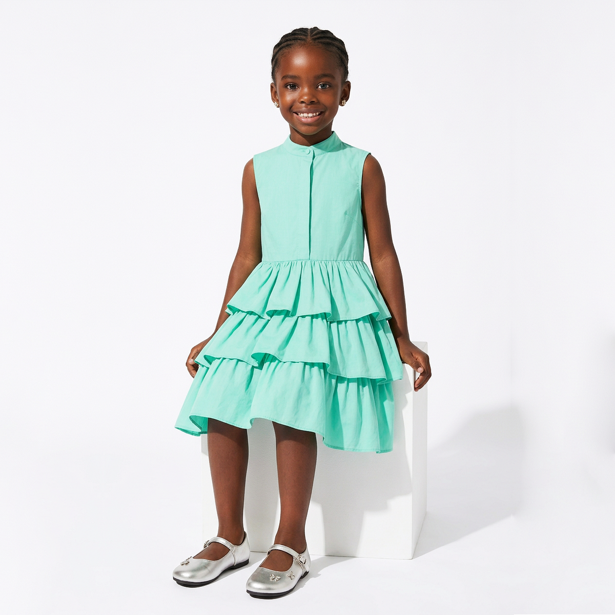 A young girl stands on a white pedestal wearing a sleeveless mint green tiered dress with a high mock neck and silver Mary Jane shoes, photographed against a clean white studio background.