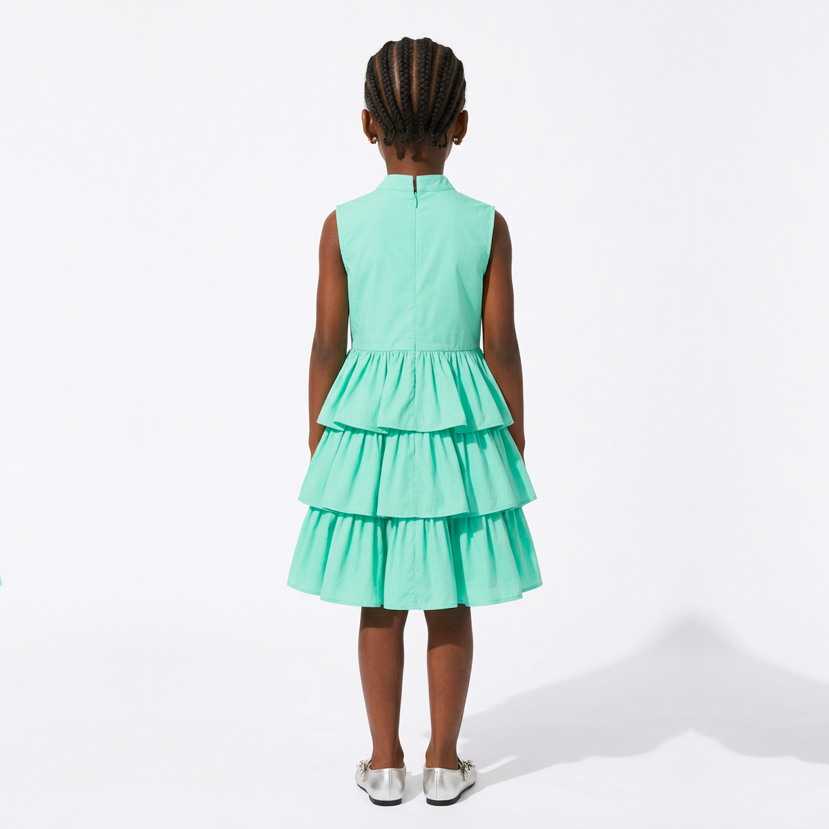 A young girl viewed from behind wearing a sleeveless mint green tiered dress and silver shoes, standing against a plain white studio background.