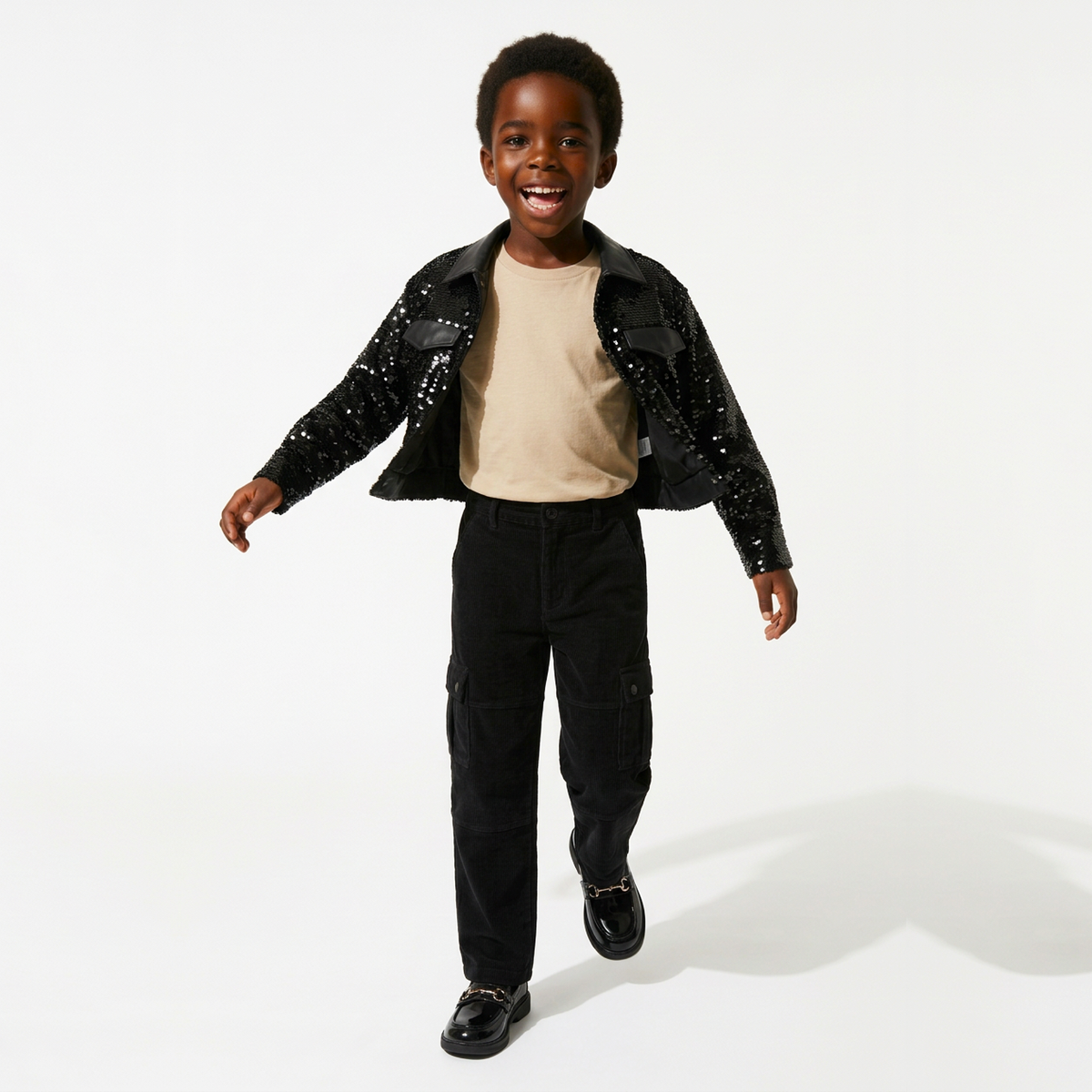 A young boy walking and smiling while wearing a black sequined jacket over a beige t-shirt, black cargo pants, and black shoes, photographed against a clean white studio background.
