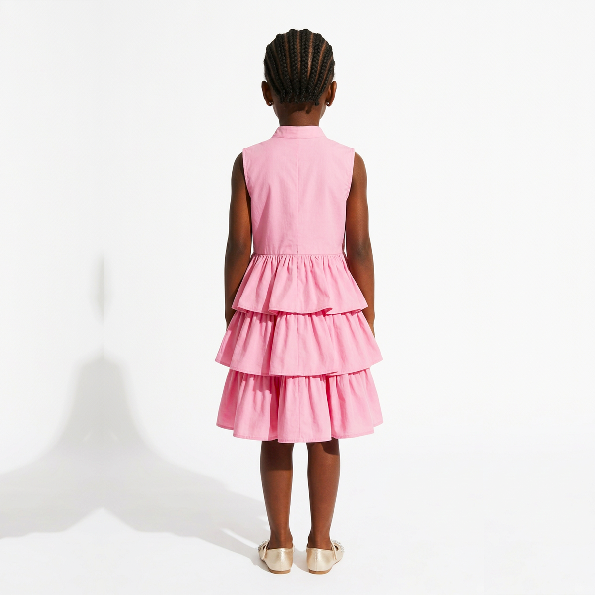 A young girl viewed from behind wearing a sleeveless pink dress with a tiered ruffle skirt, standing against a plain white studio background.
