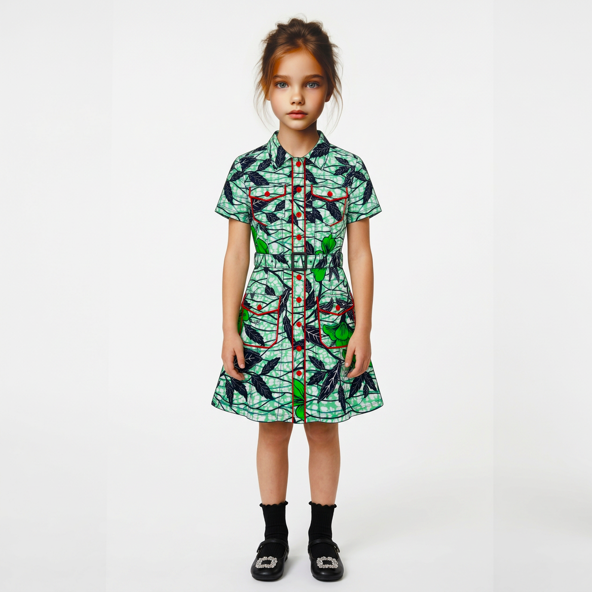 A young girl stands against a plain light gray studio background wearing a short-sleeve collared dress with an allover abstract print in green, black, white, and red, along with black socks and black shoes.