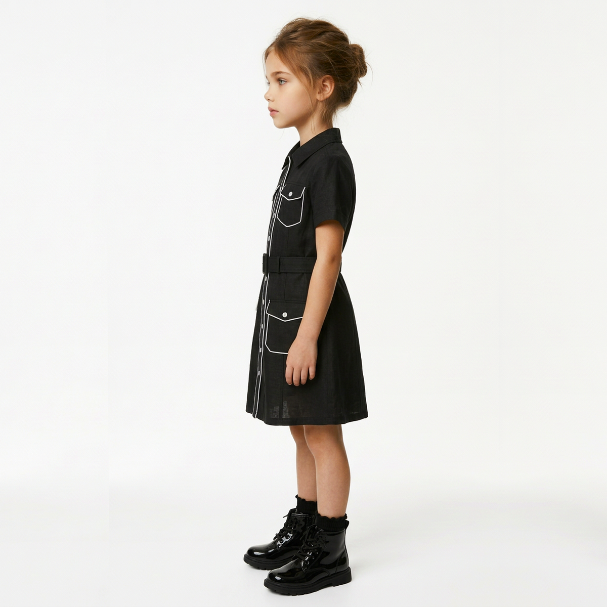 A young girl in profile wearing a black short-sleeve shirt dress with white contrast stitching and black combat boots, standing against a plain white studio background.