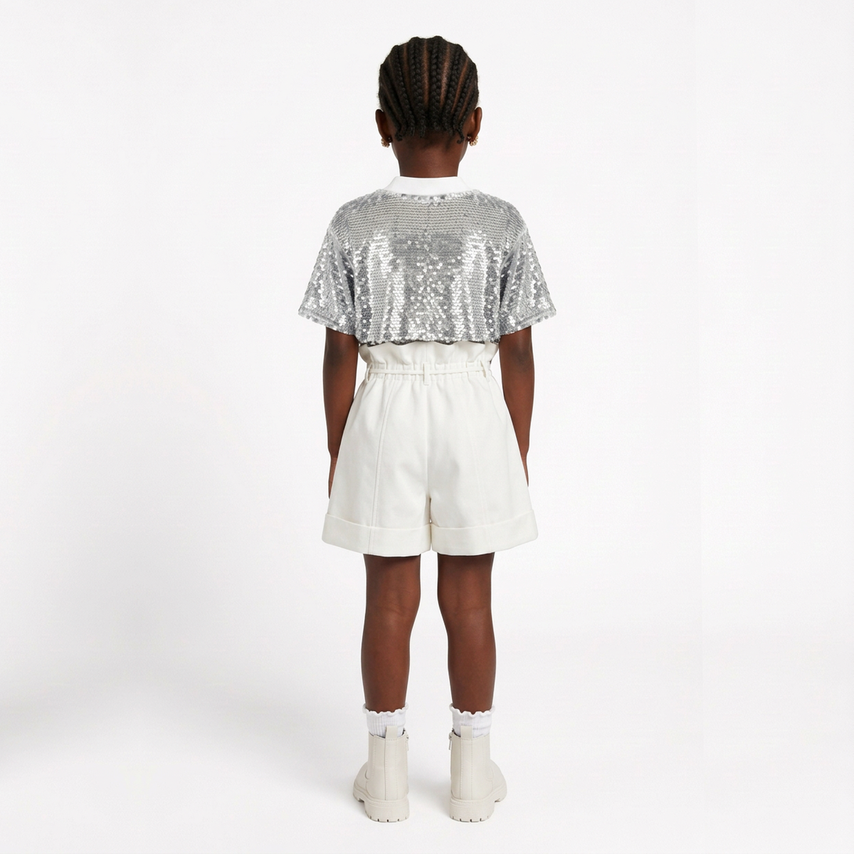 A child model shown from the back wearing a short-sleeve sequined top with a silver back and white collar, paired with off-white shorts and white boots, against a plain white studio background.