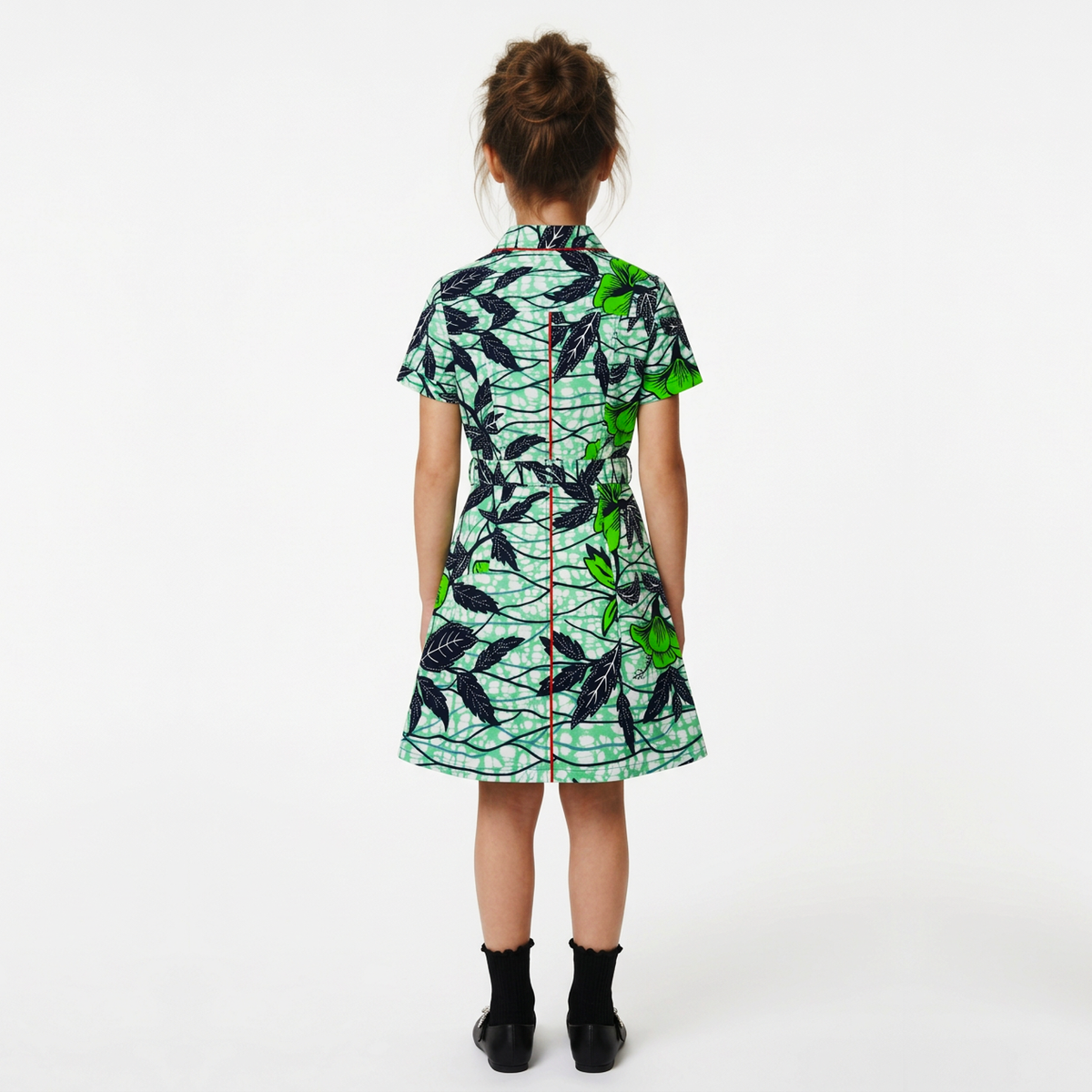 A child model viewed from the back wearing a short-sleeve collared dress with a green, black, and white leaf print, paired with black socks and black shoes, against a plain white studio background.