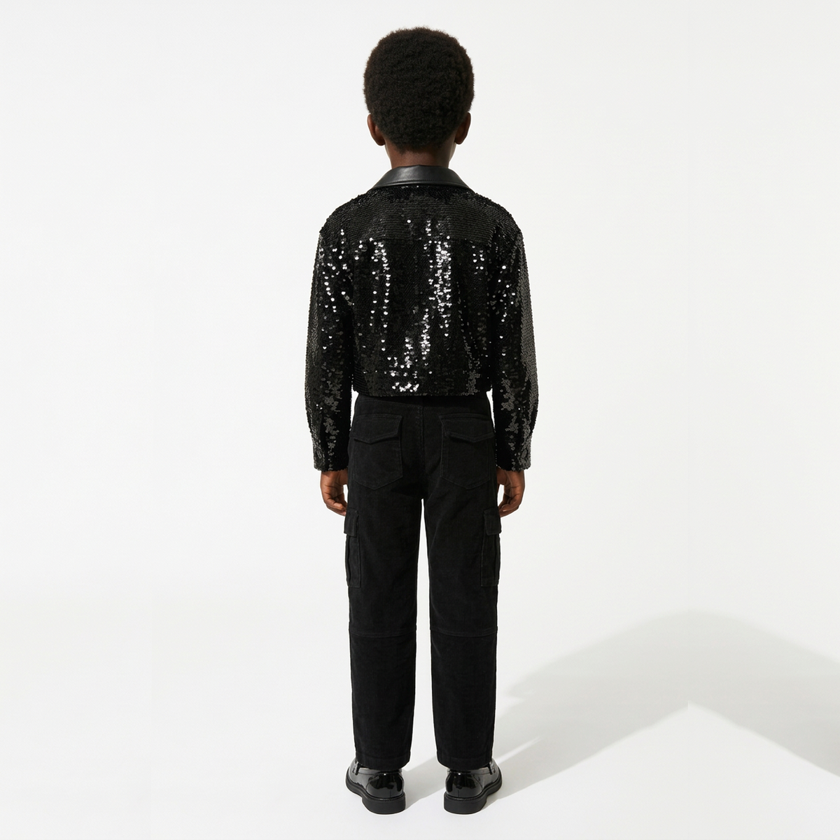 A child model shown from the back wearing a black sequined jacket with a smooth black collar, paired with black pants and black shoes, against a plain white studio background.